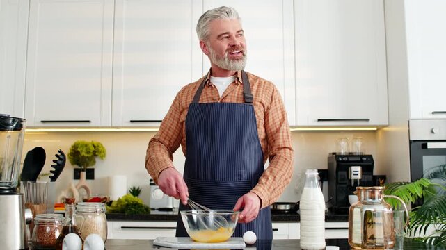 Middle-aged man whisking raw eggs in glass bowl for healthy breakfast omelet meal prep at home kitchen. Mature guy smiles while mixing for diet nutrition and wellness in modern indoors cooking space.