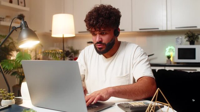Arabian man at home wears headset answers hotline call types notes opens ticket gives info. Egyptian guy sitting at table listens repeats details confirms request records data schedules follow up