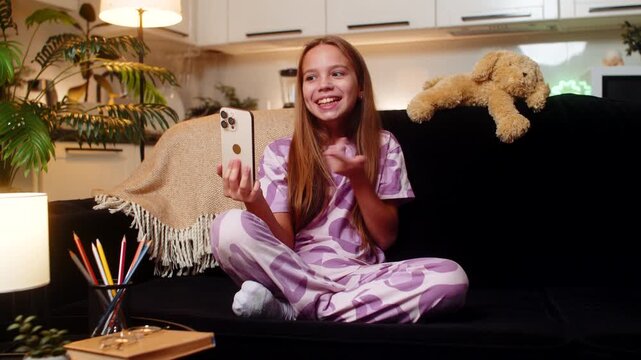Teenager girl joins video call on smartphone at night home tells school story with active gesture. Caucasian child kid on sofa at evening smiles waves hand talks voice shares homework topic in chat