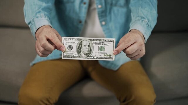 Man presents a one hundred dollar banknote directly toward the camera while sitting at home. Close-up cash concept representing personal finance, income, savings, payment, wealth, and monetary value.