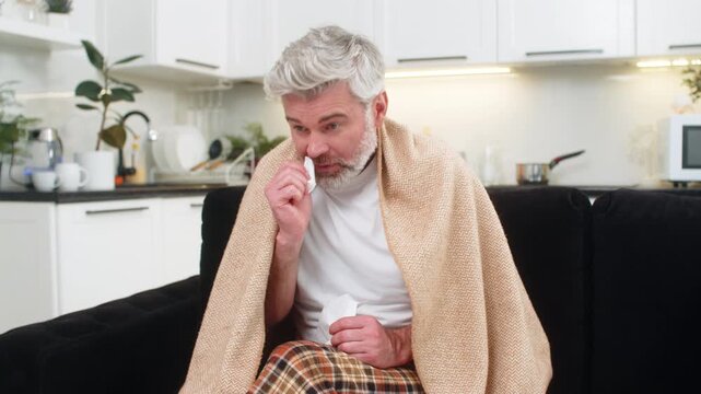 Middle-aged man at home wrapped in plaid sneezes uses tissue sniffles, wipes nose feels weak, ill. Mature guy on sofa fever chills cough fatigue breath heavy rests silent room medicine near calm wait
