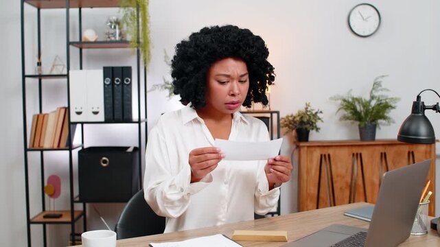African American businesswoman at home office opens envelope sees rejection letter deeply frowns grips heart. Freelancer girl at table sighs crushed planning next move vowing growth despite setback.
