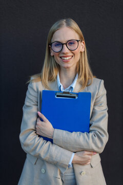 Young blonde businesswoman with glasses smiles while holding a blue clipboard 