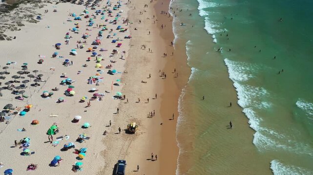 Many people relax on Praia da Rocha with umbrellas, towels, and playing in the sand aerial view. Children run, and waves crash along the shore on a sunny day.