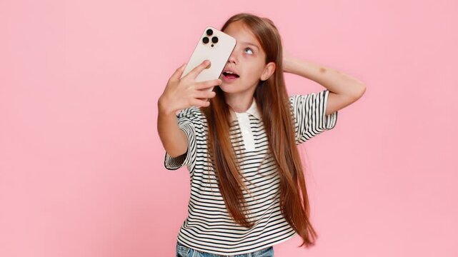 Young child girl cheerfully takes funny selfies on smartphone, joyfully laughing, posing playfully, making silly faces gestures into camera, clearly having a great time. School kid on pink background