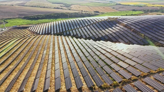 Multiple rows of solar panels stretch across a large field. The panels are aligned neatly, capturing sunlight. Green fields and crop areas surround the solar farm.