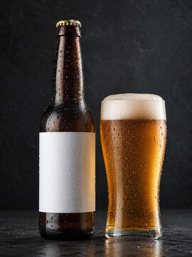 Cold craft beer bottle with blank white label and glass of lager with foam on a dark background. Beer bottle mockup for branding and packaging design.
