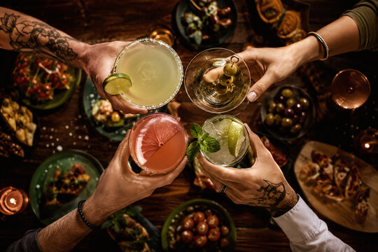 Top view of diverse friends clinking cocktails over a rustic wooden table with various appetizers and tapas. Social gathering, celebration and nightlife lifestyle concept