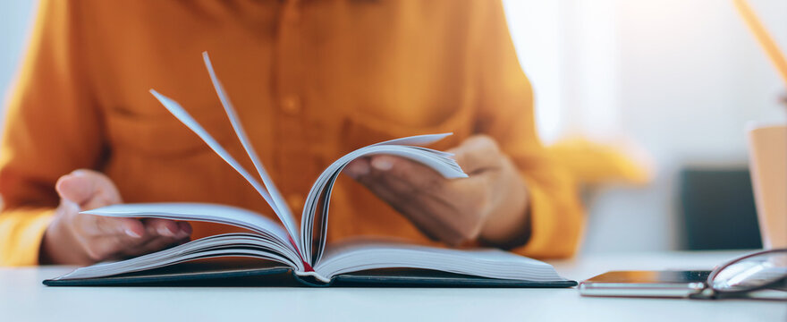Focused Unrecognizable Person Engaged In Reading Book While Sitting At A Desk In A Modern Office Environment, Surrounded By Natural Light. Concentration And Learning Concept.
