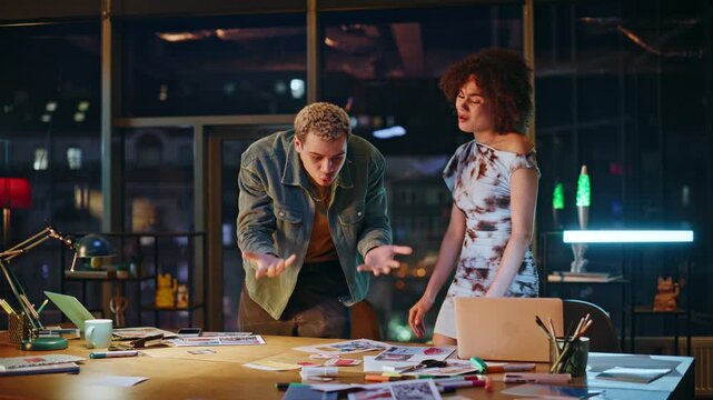 Colleagues arguing creative office night. Frustrated coworkers conflict at desk.