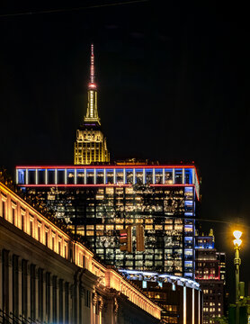 New York, NY - US - Jul 20, 2026 Night view of the Empire State Building above Midtown Manhattan, framed by a modern glass tower with red and blue lights, glowing streets and reflections
