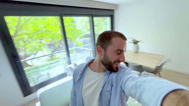 Young handsome man filming himself for his social media channel, showing his followers his new contemporary apartment as he walks around and finally sits down on the comfortable sofa