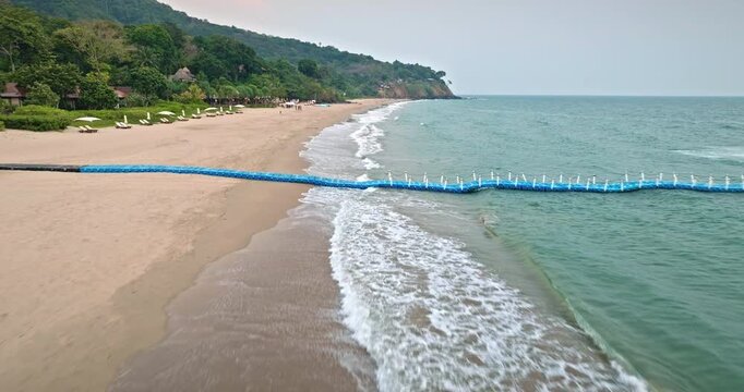 Koh Lanta tropical beach with blue floating pier extending into calm, turquoise ocean water, gentle waves breaking near the shore, lush green hillside dotted with bungalows in background. Drone flight