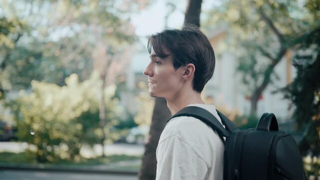 Young man in white shirt walking with backpack on tree-lined city street during sunny commute