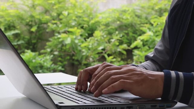 Person using laptop on table in garden at home. Close-up hands of man typing on keyboard computer. Businessman working and remote work to support customer service. Business communication technology. 