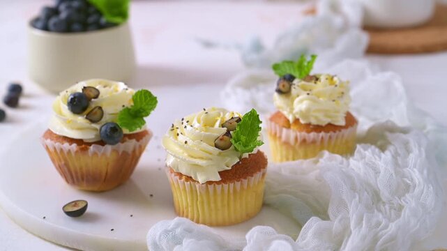 Three vanilla cupcakes featuring rich white cream frosting, fresh blueberries, and mint leaves are artfully arranged on a textured surface under bright natural light