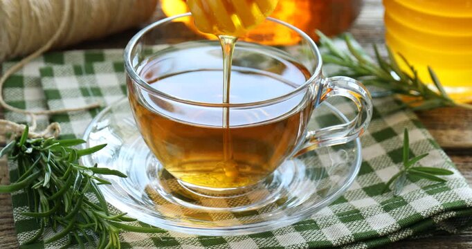 Dripping honey into cup with tea and rosemary at wooden table, closeup
