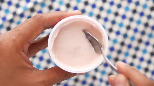 Person scooping pink strawberry yogurt with spoon from white container top view on blue patterned surface