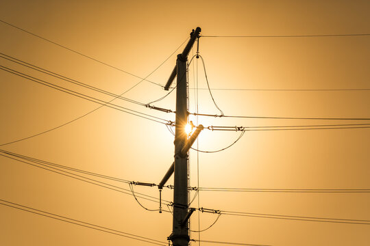 A power transmission tower stands as a silhouette against a warm sunset sky, with the sun partially hidden behind its structure.