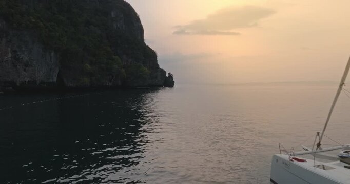 Catamaran vessel sailing in calm sea with limestone cliffs in the background during golden hour sunset, serene travel destination and luxury lifestyle experience in Thailand. Drone flight footage