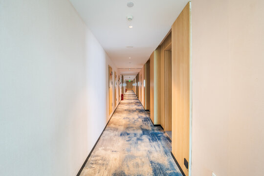 A long, empty indoor corridor with light walls, wooden doors, and patterned carpet leading to a distant exit sign.