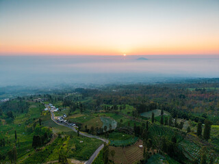 Obraz premium Aerial view of sunrise over misty valley with winding roads and scattered homes. Green fields, trees, and distant hills under soft pastel sky