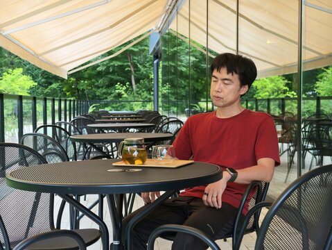 Man sits calmly at outdoor cafe table. He gazes thoughtfully toward the greenery beyond. Soft sunlight glows through the canopy above. Dark metal table holds glass of tea and napkin