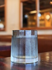Glass of sparkling water rests on a dark wooden table. Bubbles rise slowly, catching soft ambient light. Background hints at a cozy, warmly lit cafe. Image conveys freshness and quiet relaxation © Masakazu Tokashiki