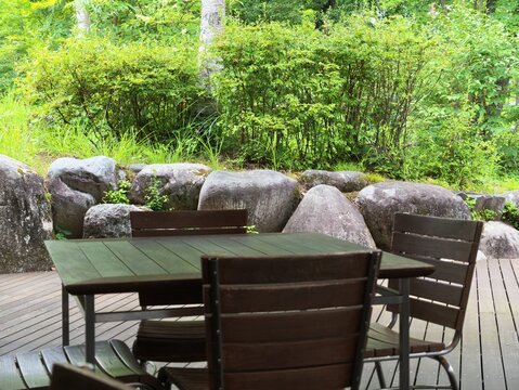Wooden table and chairs sit on a deck. Rocks and greenery frame the tranquil garden space. Natural light enhances the peaceful, rustic feel. Ideal for outdoor dining or quiet relaxation scenes