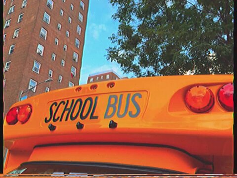 Close-up view of the back of a yellow school bus, with a nostalgic vhs aesthetic, parked on an urban street next to a brick building and trees, symbolizing public education