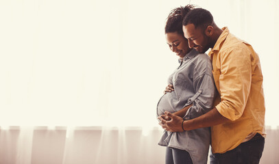 Fototapeta premium Joyful African American Couple Smiling While Embracing Pregnant Belly In Bright Room Filled With Natural Light. Happy Expecting Parents Enjoying Intimate Moment Together In Family Setting.