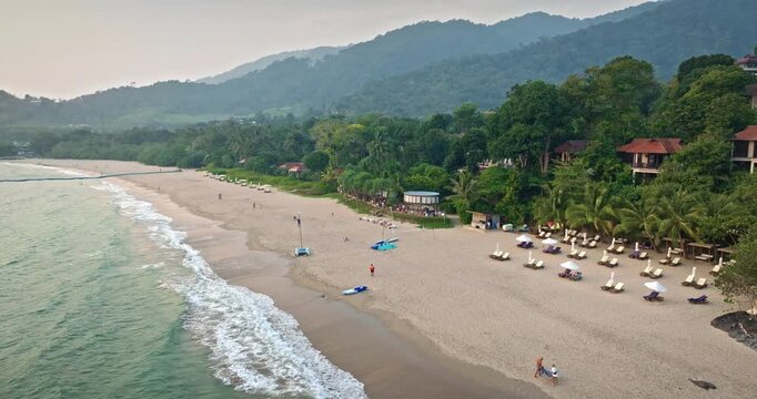 Koh Lanta Island aerial seascape. Peaceful Thailand beach with people relaxing on white sand, enjoying sun loungers and umbrellas, next to a lush green hillside resort. Drone flight panorama