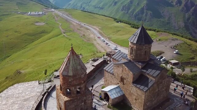 Aerial view of Gergeti trinity church perched on high mountain slope in Kazbegi Georgia with tourists exploring historic landscape.