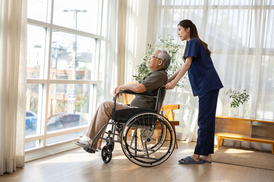 Caring female nurse assisting senior man in wheelchair looking through large bright window with calm peaceful expression inside comfortable modern nursing home facility living room