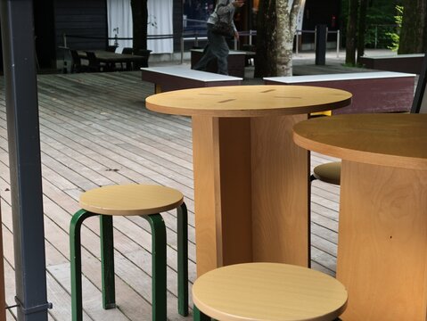 Wooden tables and stools sit on a deck. Green metal legs support simple, round seating. Person walks past, unnoticed by the furniture. Urban space blends casual, minimalist design