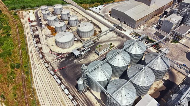 Large metal storage tanks and silos are seen at an industrial site. Trains are parked nearby. The sun is shining and activity can be observed in the area.