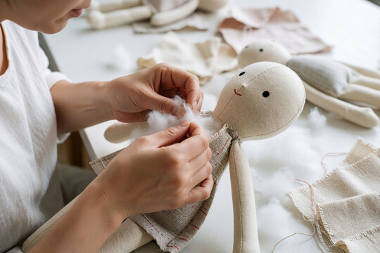 Tailor fills handmade fabric doll with soft cotton fiber at workshop table. Woman sews textile stuffed toy for children in small creative atelier. Artisan crafts cuddly present using needle, thread.