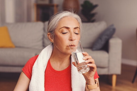 Focused Senior Woman Hydrating By Drinking Water After Workout At Home, Sitting In Living Room With Towel Around Neck, Promoting Wellness And Fitness Lifestyle.