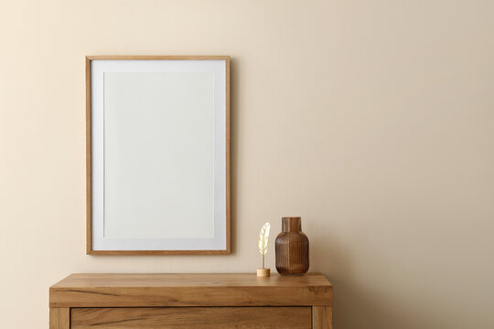 Vertical wooden frame with blank white poster mockup on a light beige wall above a rustic wooden dresser, a rustic wooden dresser sits below the frame
