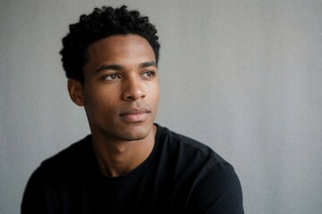 Obraz premium Young black man with curly hair looking away on grey studio background. Serious handsome guy wears casual black shirt posing for portrait. Confident male model with thoughtful facial expression poses