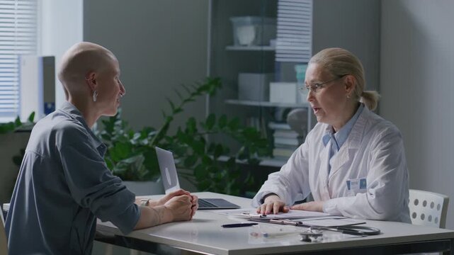 Bald female oncology patient smiling and shaking hands with supportive doctor at desk during consultation in medical office