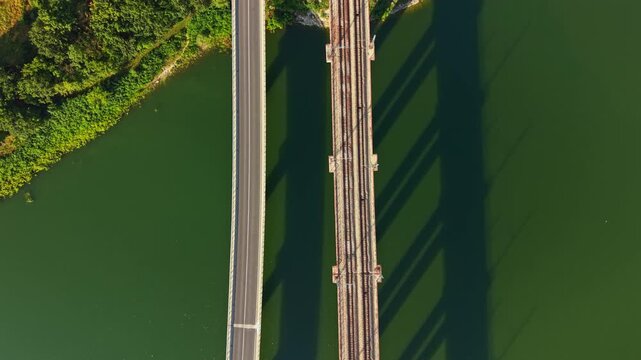 Overhead aerial view shows two parallel bridges crossing green water, with road traffic on one span and railway tracks on other, plus single vehicle moving steadily beside calm reflective water