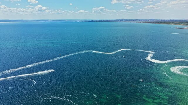 Participants race jet skis at high speed on clear waters under blue skies. The scene shows waves created by the jet skis and a distant shoreline.