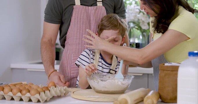 Caucasian mother gently wipes flour from young son's face while boy keeps stirring batter, as father stands nearby during home baking lesson with caring parenting, teamwork and family bonding.