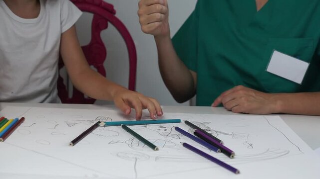 A female therapist in green uniform handing a pink pencil to a young girl seated at a desk during a pediatric care session at home.