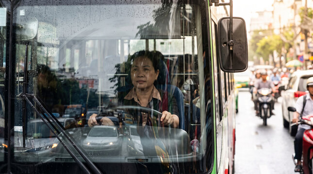 Female bus driver navigating through busy city traffic with motorbikes on a sunny day in Vietnam showing dedication and professional transit services in urban public transportation.
