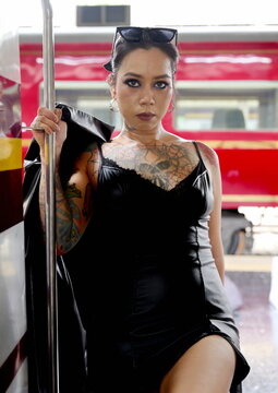 Asian woman with chest and arm tattoos wearing a black dress posing at the entrance of a train carriage