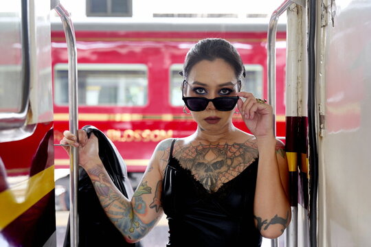 Asian woman with chest and arm tattoos wearing a black dress posing at the entrance of a train carriage