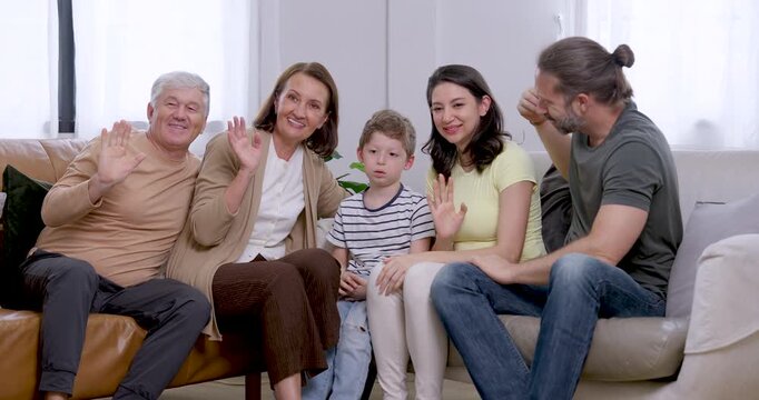 Slow motion young son between Asian mother and Caucasian father as senior grandparents smile and wave toward camera, expressing cheerful greeting and multigenerational family togetherness at home.