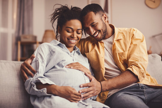 Joyful African American Couple Smiling At Each Other While Enjoying Time Together In Cozy Living Room, Embracing Pregnant Belly In Maternity Celebration. Pregnancy And Family Concept.